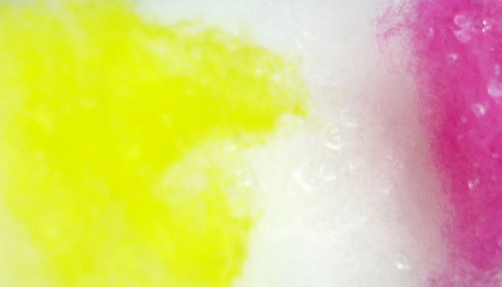 Ein Bild von Wollfasern: Links gelb, in der Mitte weiß, rechts rosa.