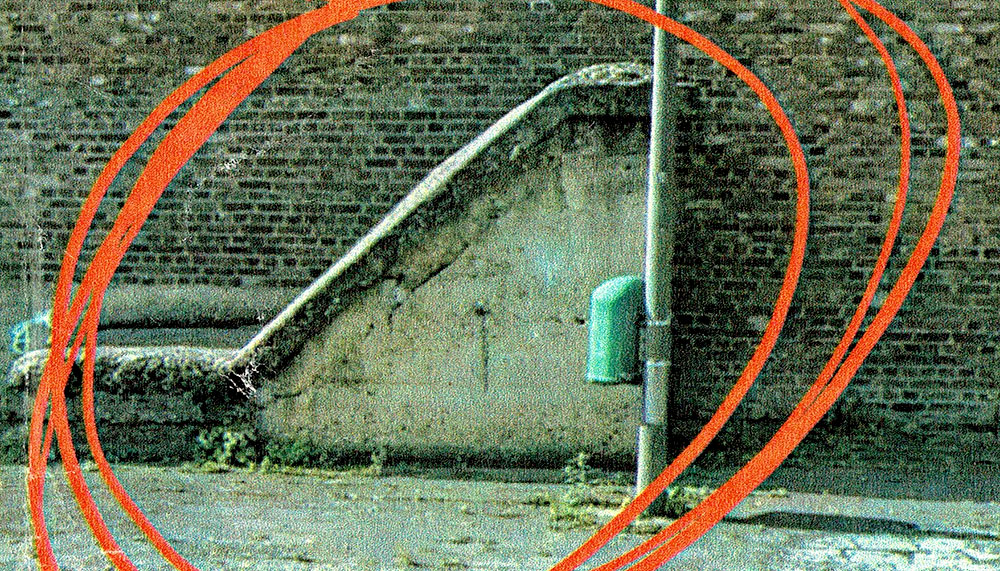 Foto von Backsteingebäude-Wand. Davor rot eingekreist ein Betonvorsprung. Links davon ein Mülleimer.