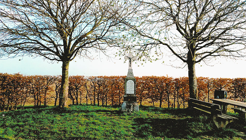 Foto eines Steinkreuzes, dahinter eine herbstliche Hecke und Felder. Rechts vom Kreuz ein Picknicktisch mit Bänken.